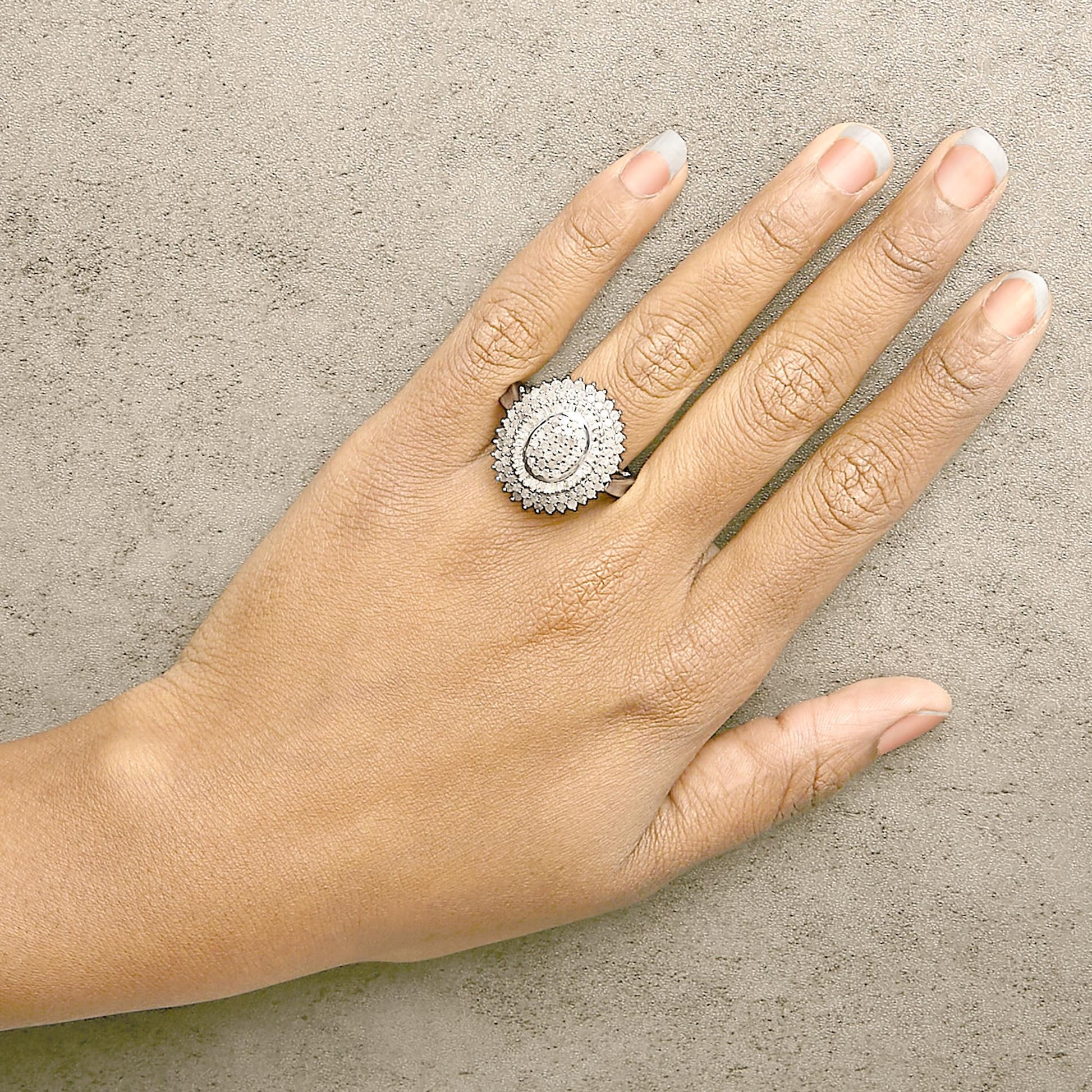 White metal ring with an oval cluster of round white gemstones in prong settings surrounded by baguette white gemstones in channel settings, shown worn on a model's finger from a top angle.