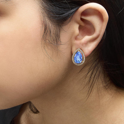 Close-up side view of a model wearing a yellow metal earring featuring a pear-shaped blue gemstone surrounded by round white gemstones in a prong setting.