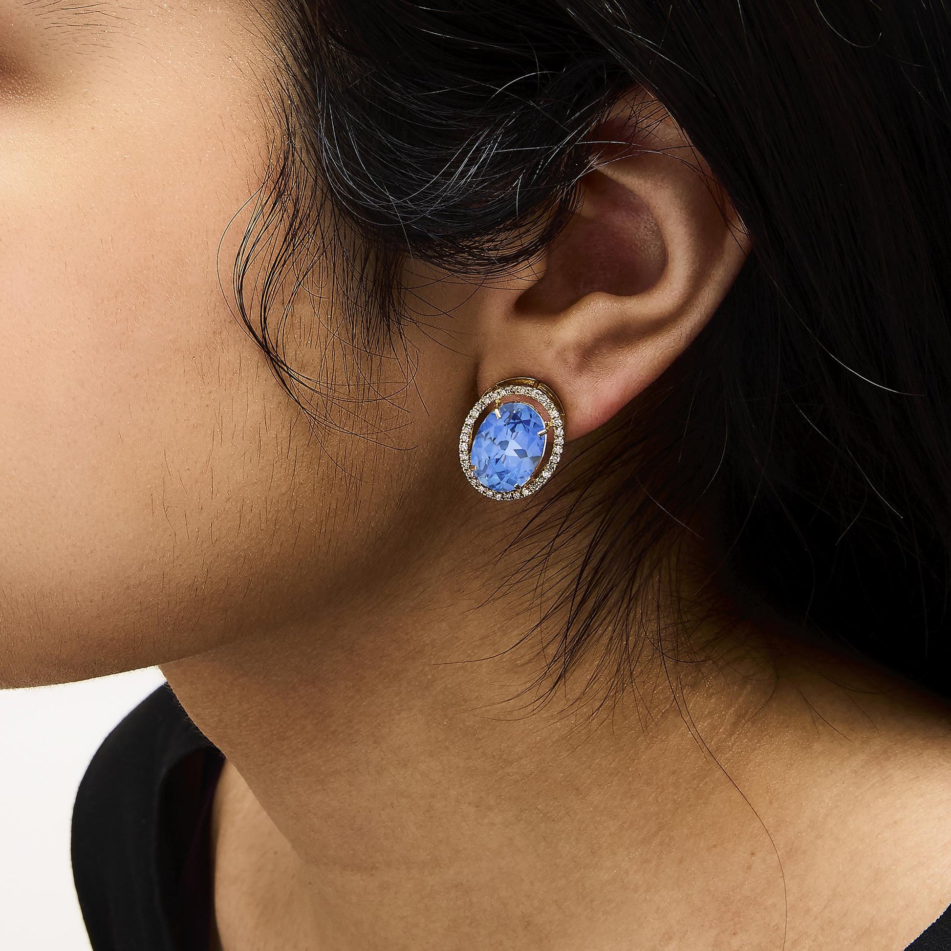 Close-up side angle of a model wearing yellow metal earrings featuring an oval blue gemstone in the center surrounded by a halo of small round white gemstones in prong settings.