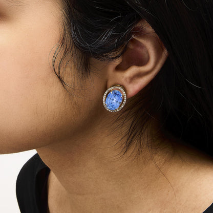 Close-up side angle of a model wearing yellow metal earrings featuring an oval blue gemstone in the center surrounded by a halo of small round white gemstones in prong settings.
