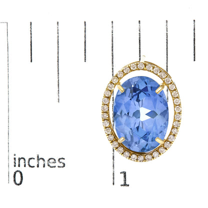 Close-up image of a yellow metal earring featuring an oval blue gemstone in a four-prong setting, surrounded by a halo of small round white gemstones in prong settings, shown on a scale with inch markings, shot from a top angle.
