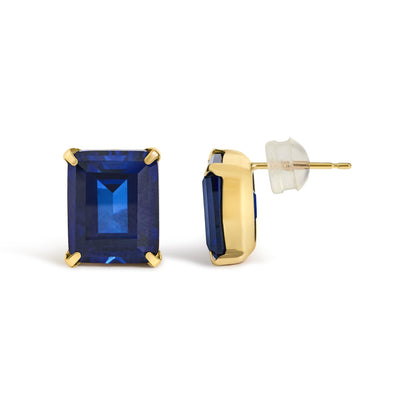 Close-up front and side view of yellow metal stud earrings featuring blue emerald-shaped gemstones in four-prong settings with clear backs, shown on a white background.