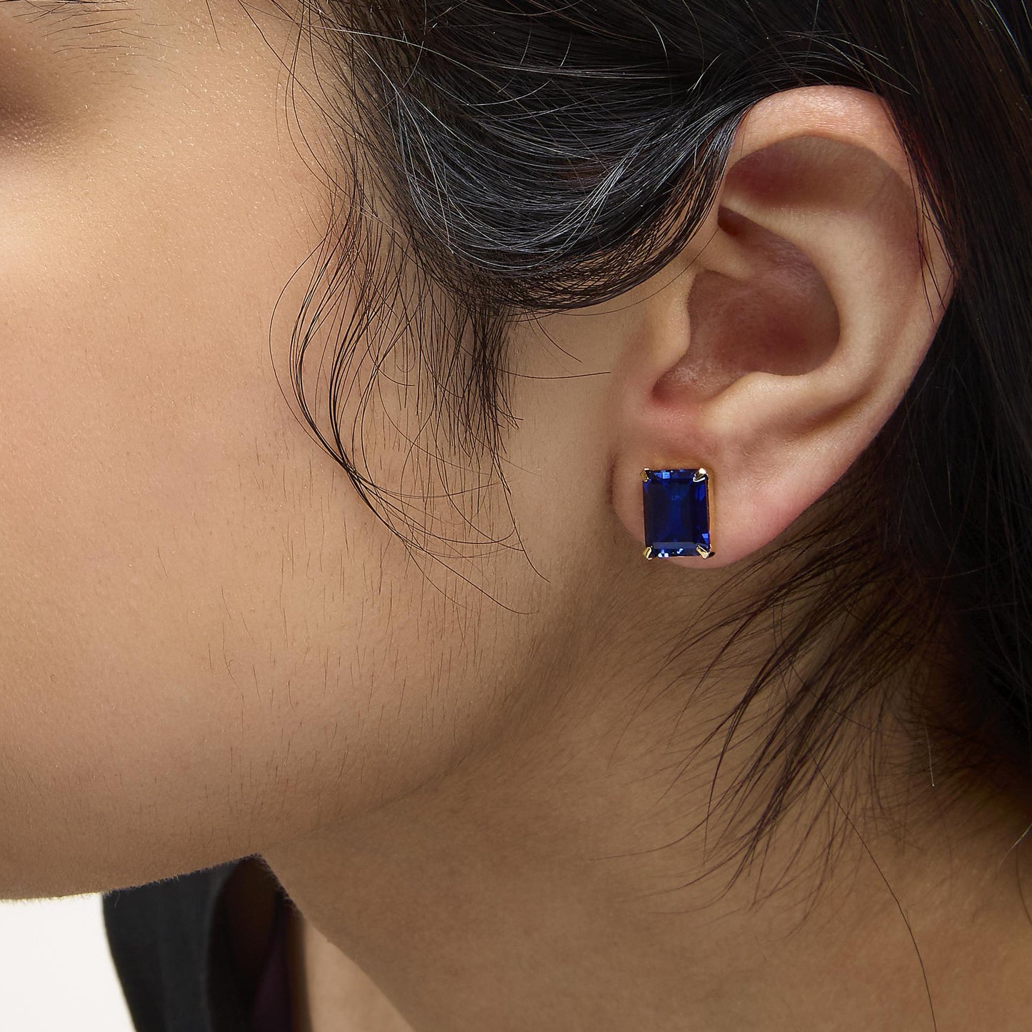 Close-up side view of a model wearing rectangular blue gemstone stud earrings set in yellow metal with 4-prong settings.
