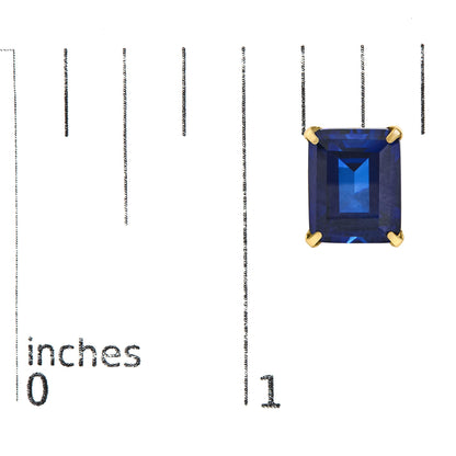 Close-up image of a yellow metal earring with a large emerald-shaped blue gemstone in a four-prong setting, shown on a measurement scale from top front angle.