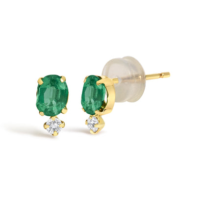 Close-up angled view of yellow metal earrings featuring oval green gemstones held by prongs above round white gemstones in prong settings, with clear backings shown.