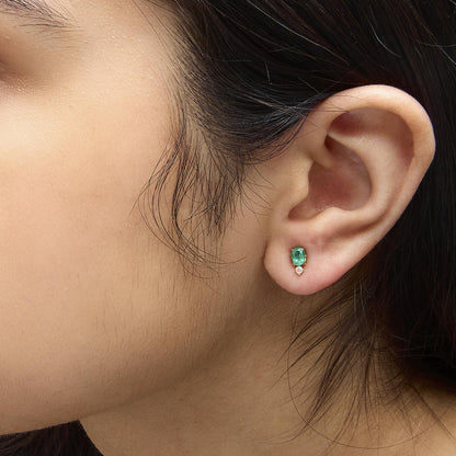 Close-up front angle of a model's ear wearing yellow metal earrings with oval green gemstones set above round white gemstones.
