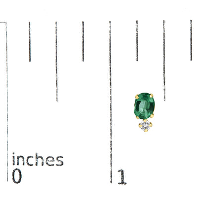Close-up front angle of a yellow metal earring featuring an oval green gemstone set above a smaller round white gemstone, both in four-prong settings, shown on a measurement scale.