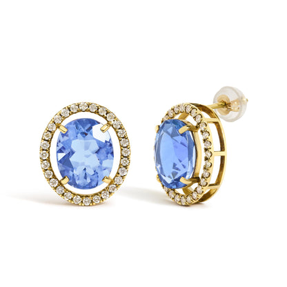 Close-up side angle of yellow metal oval earrings with large blue oval gemstones in four-prong settings, surrounded by a halo of small round white gemstones in prong settings.