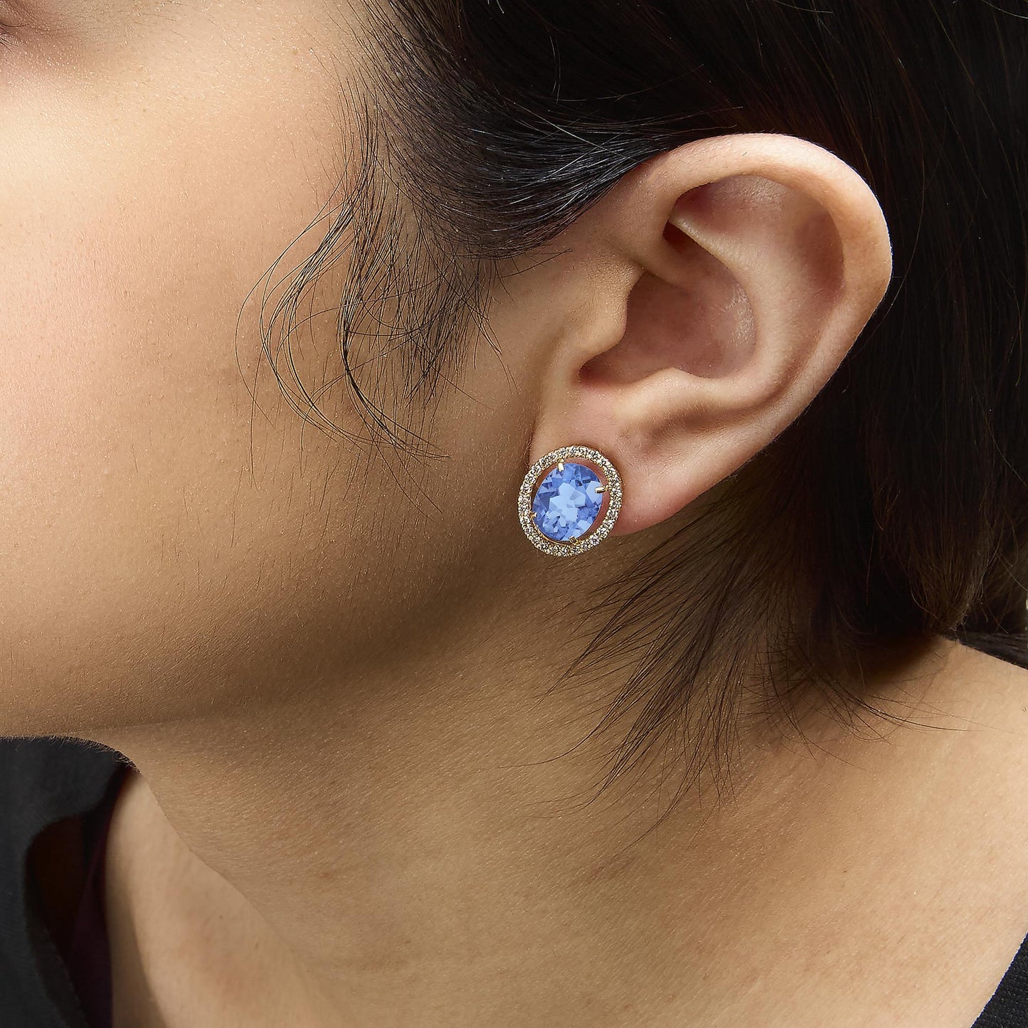 Close-up side view of a model wearing yellow metal earrings featuring a central oval blue gemstone surrounded by a halo of small round white gemstones in prong settings.
