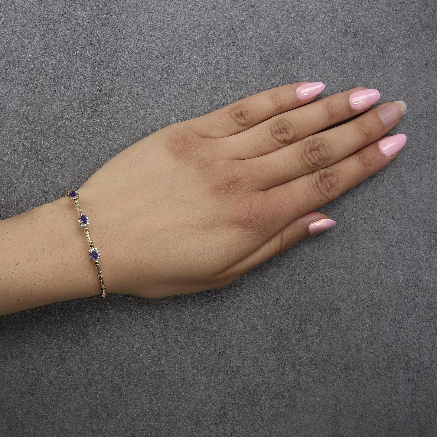 Yellow metal bracelet with oval purple gemstones and round white gemstones in prong settings, shown worn on a model's wrist in a top-down angle.