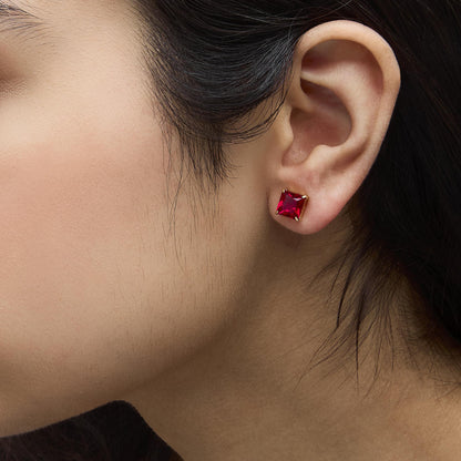 Close-up side view of a model wearing yellow metal stud earrings featuring red princess-shaped gemstones in a 4-prong setting.