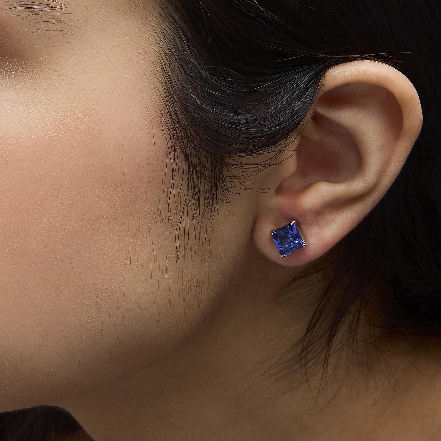 Close-up side view of a model wearing white metal stud earrings featuring princess-shaped blue gemstones in a 4-prong setting.