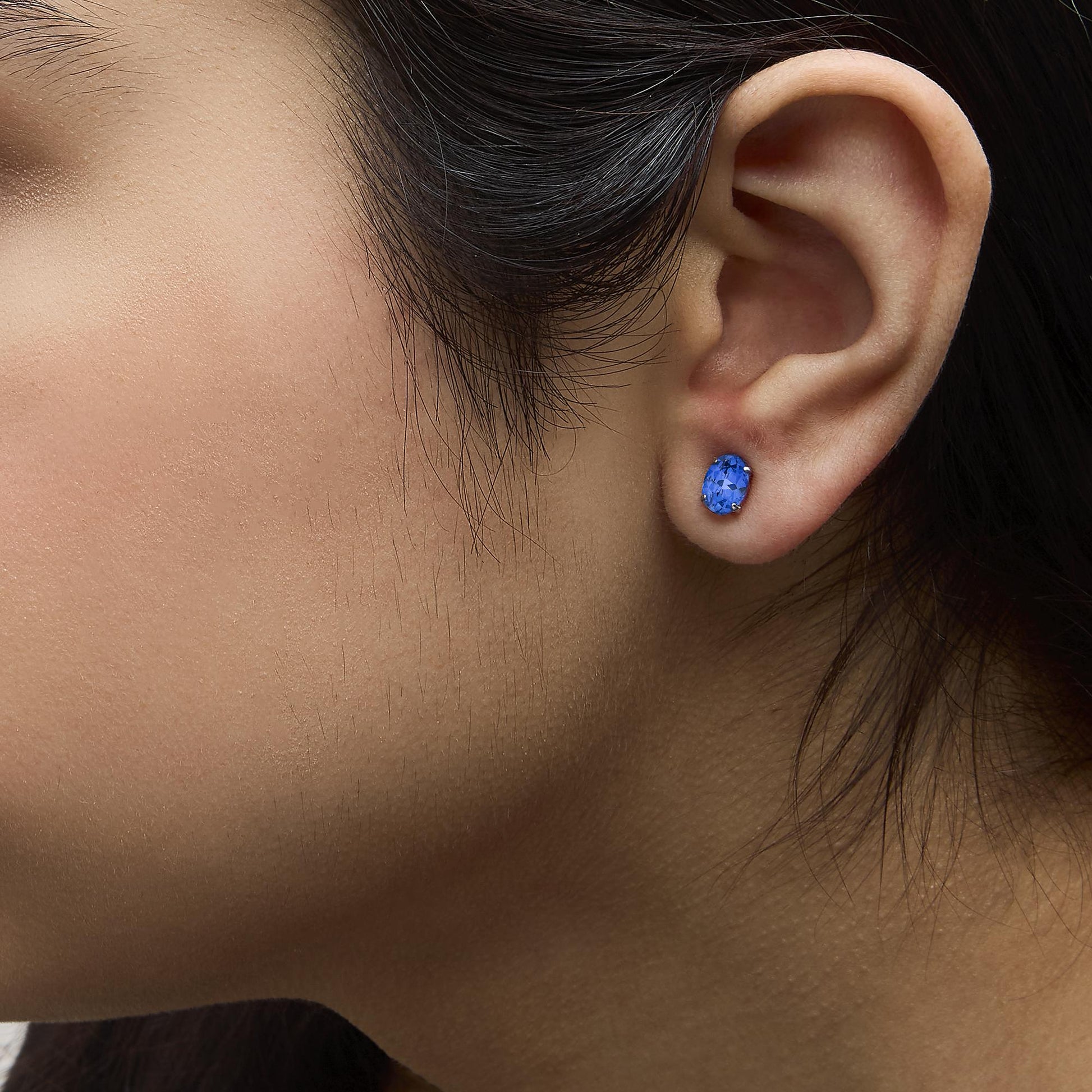 Close-up side view of a model wearing white metal stud earrings with oval-shaped blue gemstones in a four-prong setting.