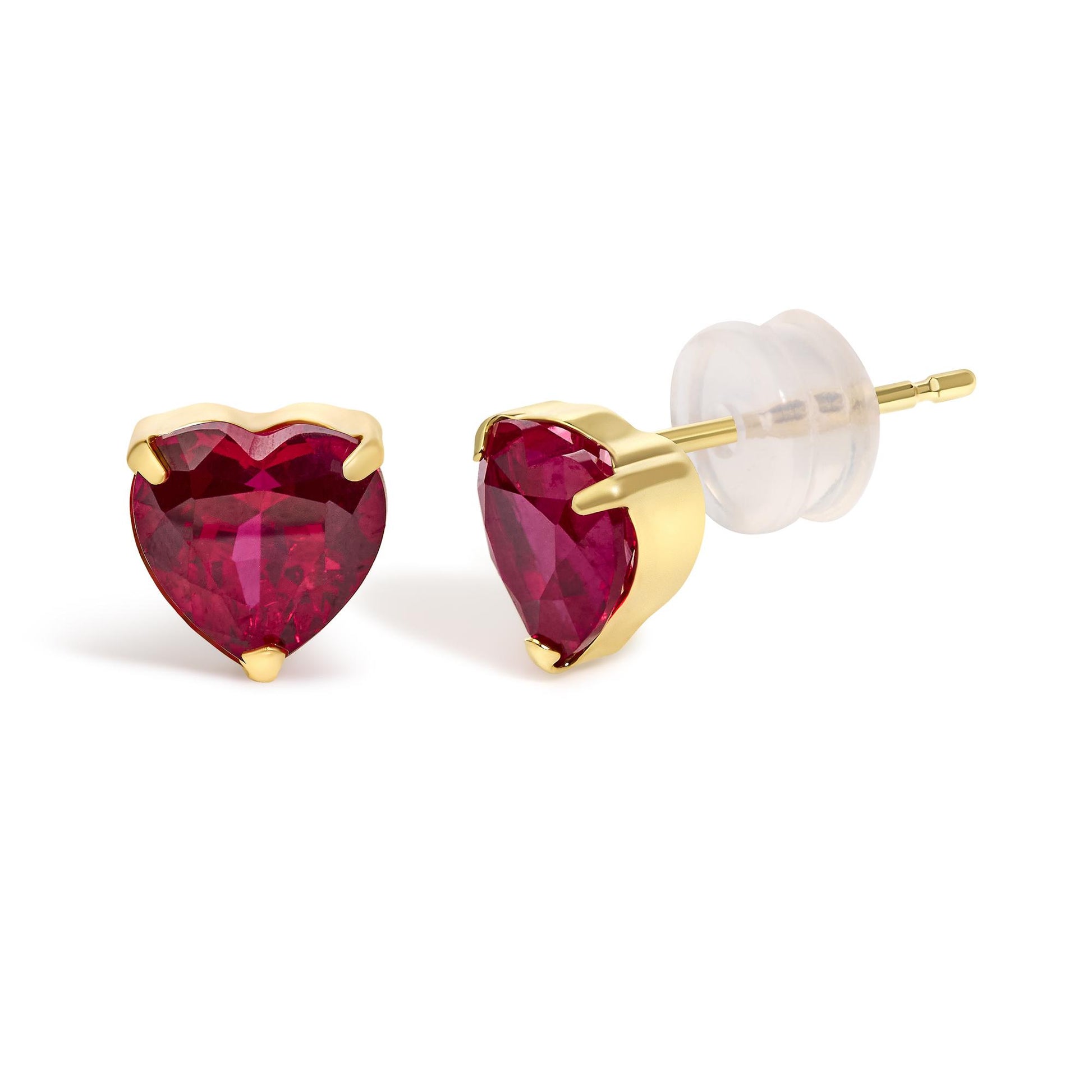 Close-up front and side angle of a pair of yellow metal stud earrings, each featuring a heart-shaped red gemstone in a three-prong setting, shown on a white background.