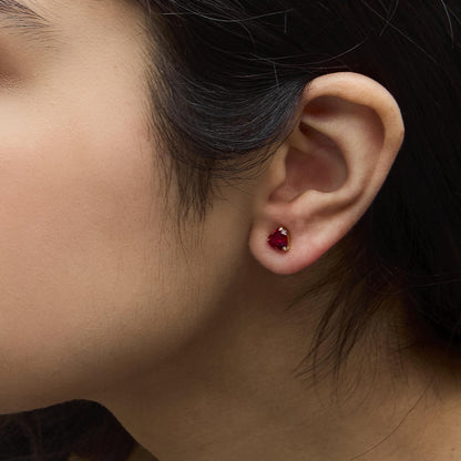 Close-up side view of a model wearing yellow metal stud earrings with heart-shaped red gemstones in a 3-prong setting.