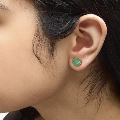 Close-up side view of model wearing yellow metal earrings with heart-shaped green gemstone in bezel setting.