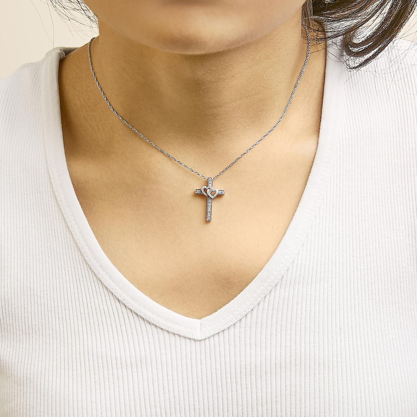 Close-up front view of a necklace worn by a model featuring a white and rose colored pendant in the shape of a cross with two heart shapes intertwined, decorated with round white gemstones in a pave setting.