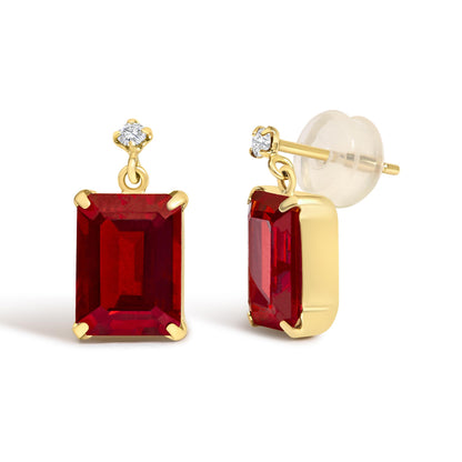 Close-up angled view of yellow metal earrings featuring a red emerald-shaped gemstone in a four-prong setting, topped with a round white gemstone in a four-prong setting, shown on a white background.