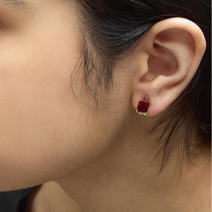 Close-up side view of a model wearing yellow metal earrings featuring a red emerald-shaped gemstone with a white round gemstone above it, both set in 4-prong settings.