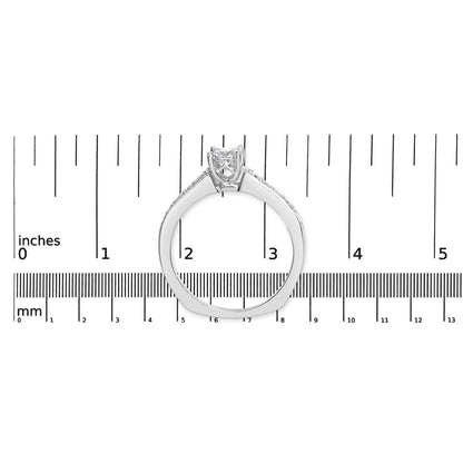 White metal ring with a princess shape gemstone in a four-prong setting flanked by round gemstones in a shared prong setting, shown from the side on a ruler for size reference.