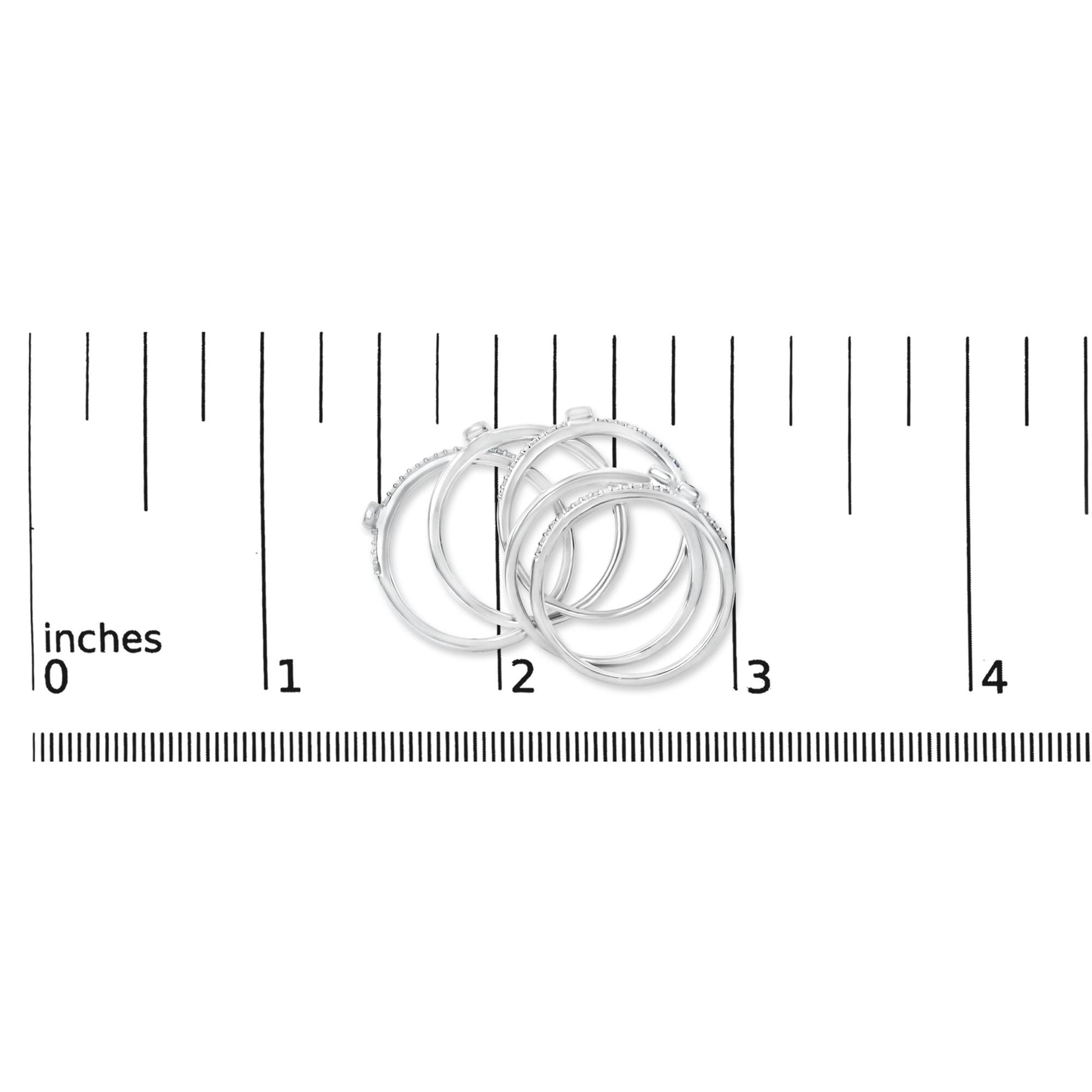 Close-up front angle of four white rings with small round white gemstones in bezel and prong settings, shown on a ruler scale.