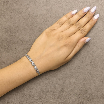 Close-up angled view of a white metal bracelet with round white gemstones in prong settings, shown worn on a model's wrist.