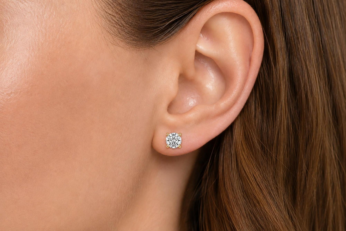Close-up side view of a model wearing round white gemstone stud earrings with four-prong yellow metal settings.