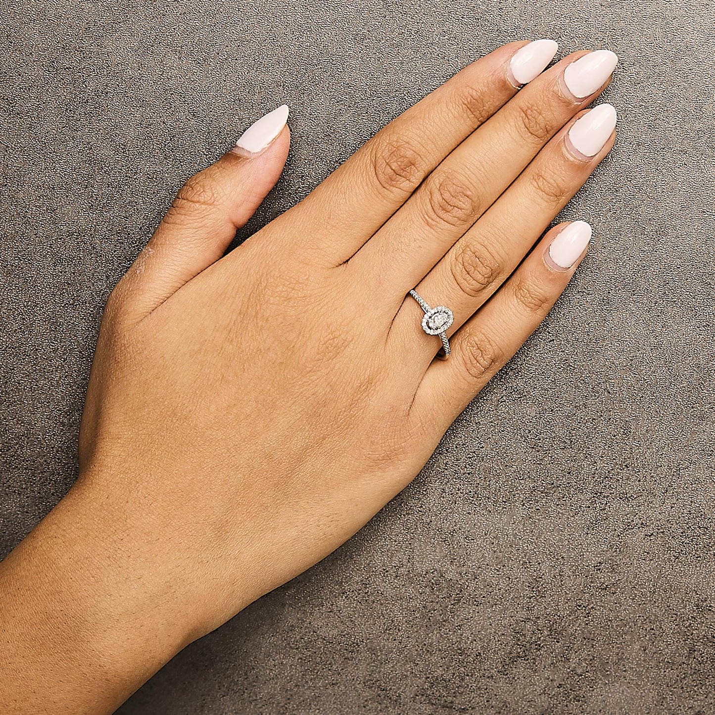 White metal ring with a central round white gemstone surrounded by smaller white gemstones in a halo setting, shown worn on a model's hand, angled from above.
