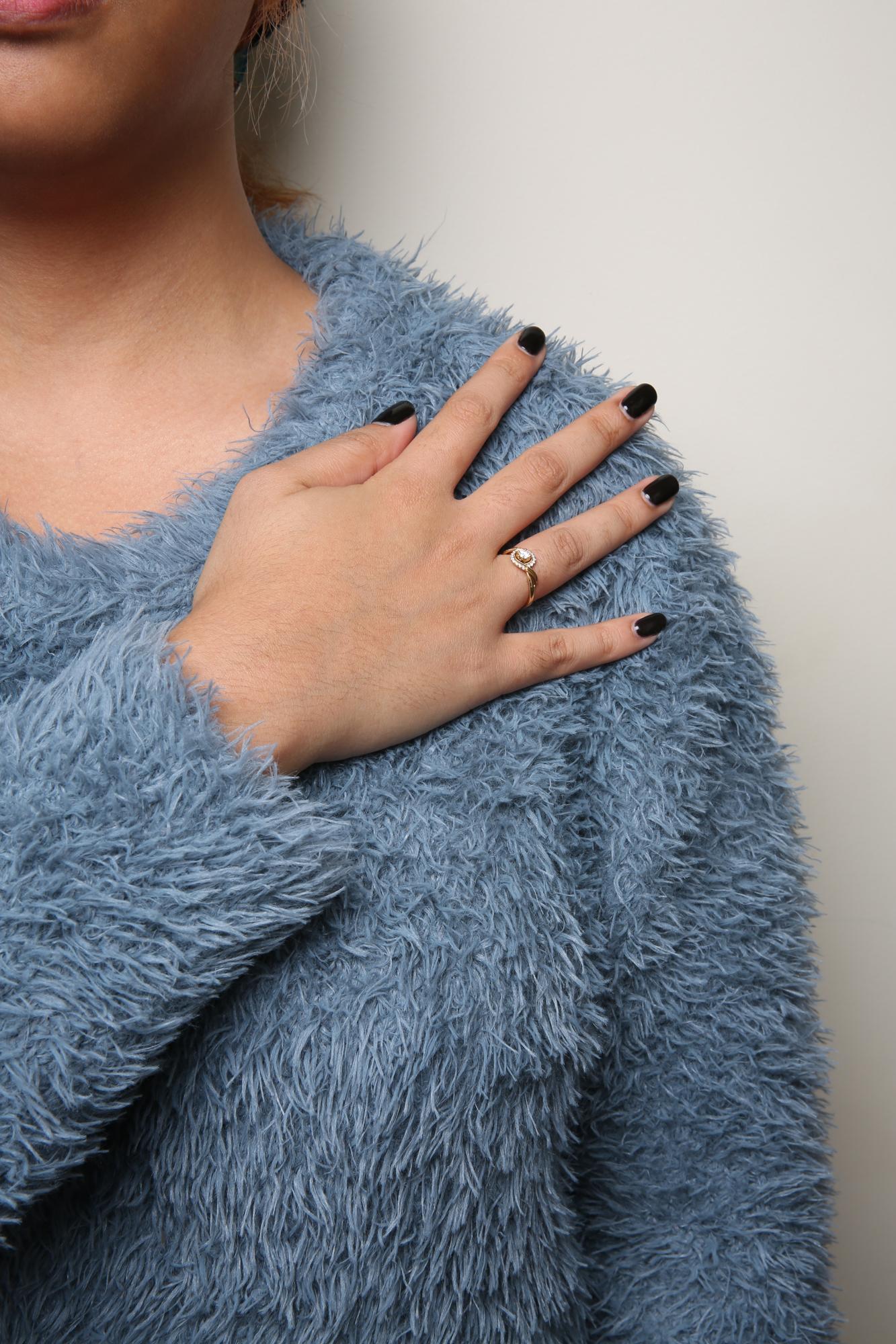 Yellow metal ring with a single round white gemstone in a 4-prong setting, surrounded by smaller round white gemstones in a shared prong setting, worn on a model's finger, shot from the front at an angle.