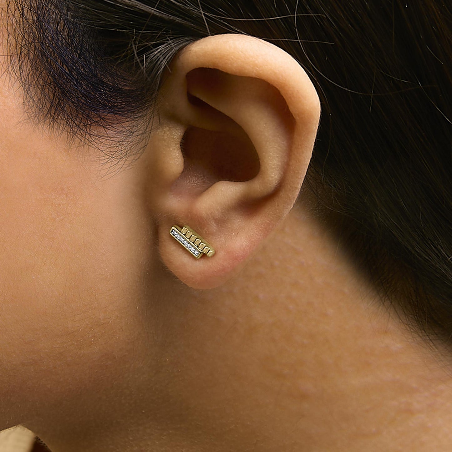 Close-up side view of a model wearing a yellow metal bar-shaped earring with a row of small round white gemstones in a pave setting.