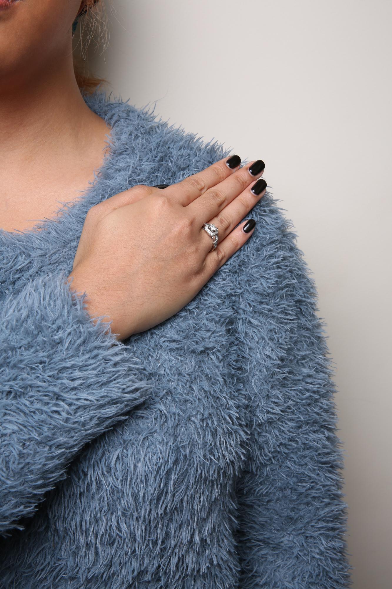Close-up of a white metal ring on a model's finger featuring one princess shape white gemstone in a four-prong setting and multiple round shape white gemstones in a shared prong setting, photographed from the side.
