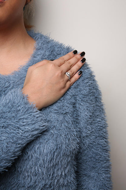 Close-up of a white metal ring on a model's finger featuring one princess shape white gemstone in a four-prong setting and multiple round shape white gemstones in a shared prong setting, photographed from the side.