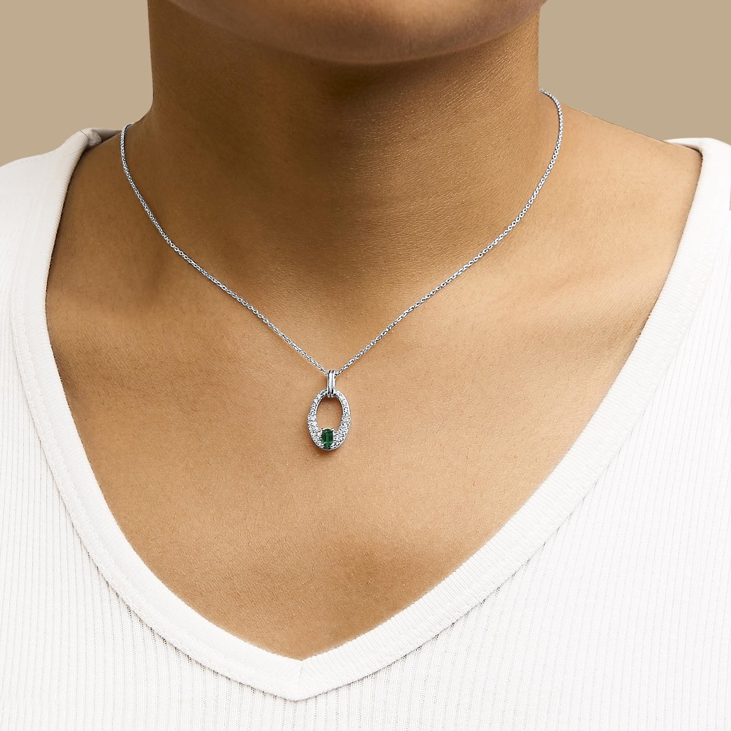 Necklace with an oval-shaped pendant shown worn on a model, featuring a white metal oval frame set with multiple small round white gemstones and a single oval green gemstone at the bottom center, photographed from the front.