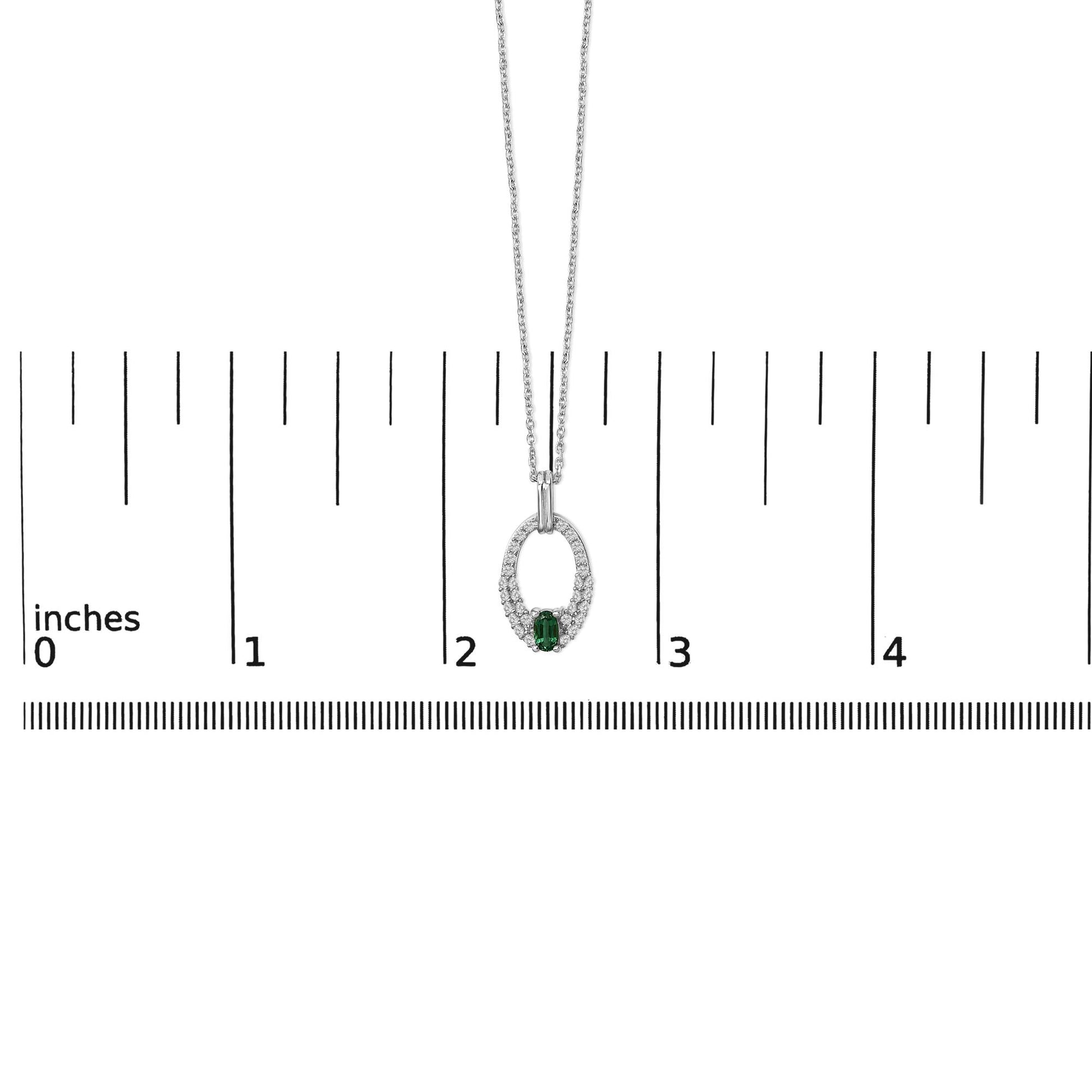 White metal necklace with an oval-shaped green gemstone center set in a prong setting, surrounded by multiple round white gemstones, shown on a ruler scale from a front angle.