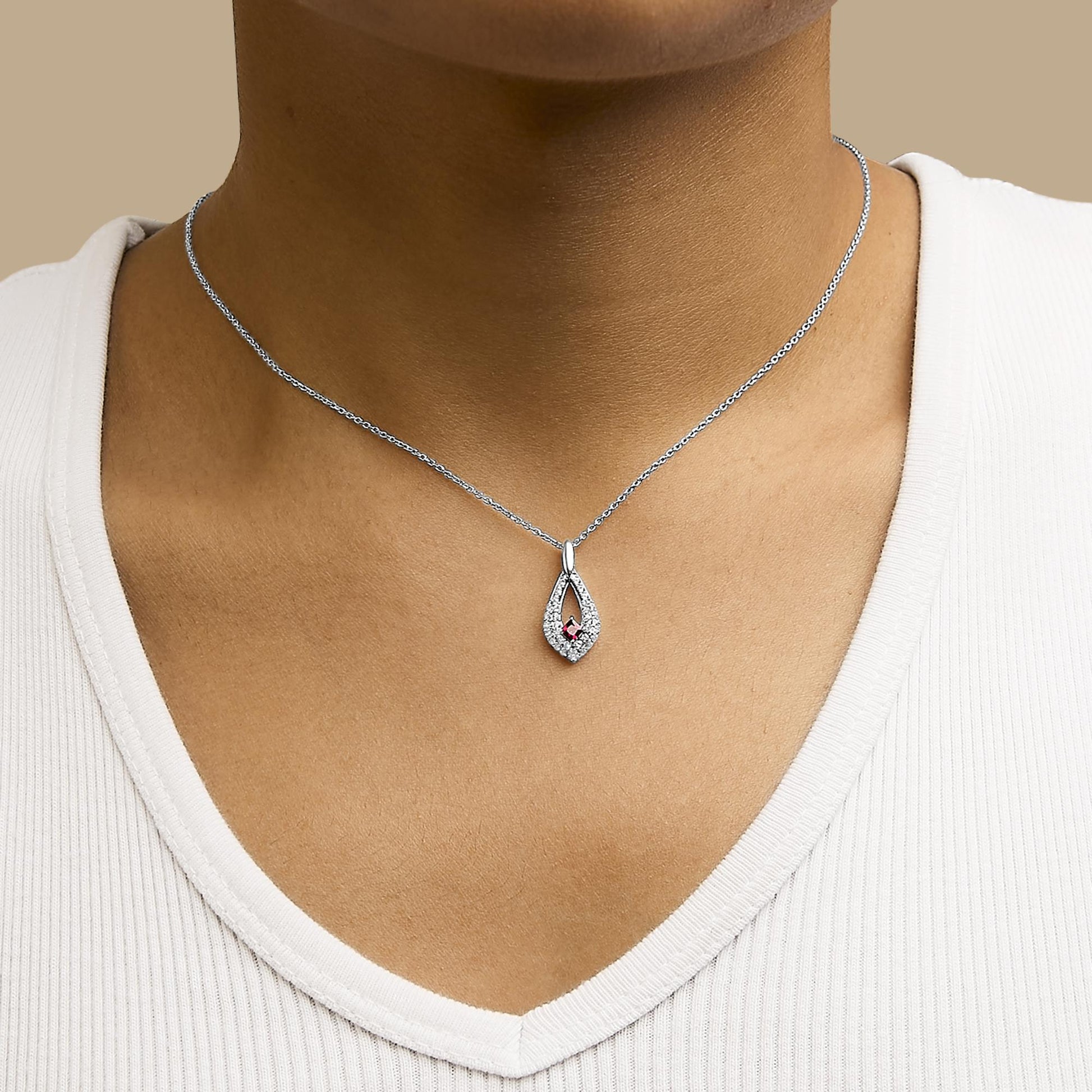 Close-up frontal view of a white metal necklace with a teardrop-shaped pendant shown worn on a model. The pendant features a central red princess-shaped gemstone surrounded by small round white gemstones.