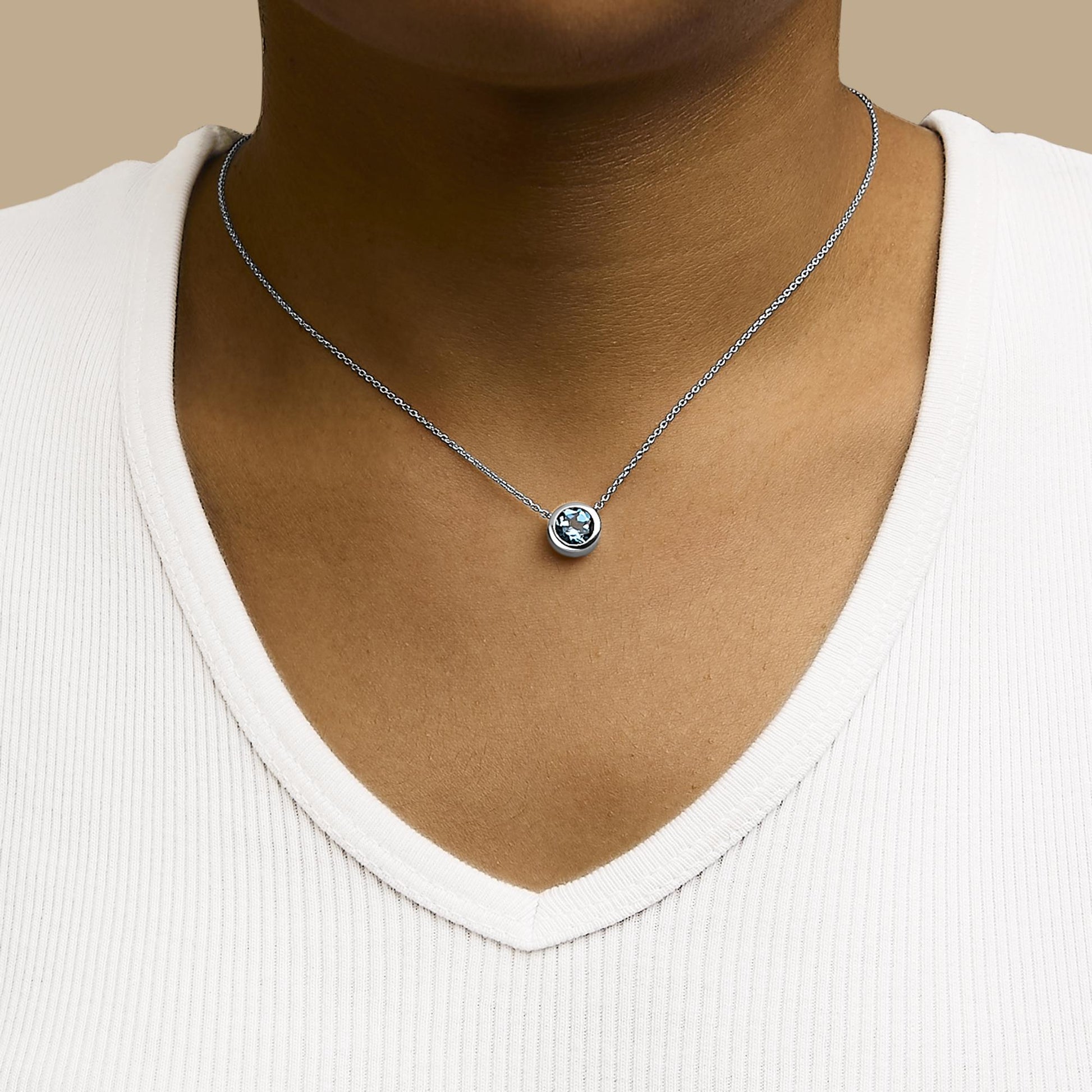 Close-up front view of a white necklace with a round blue gemstone in a bezel setting, shown worn by a model.