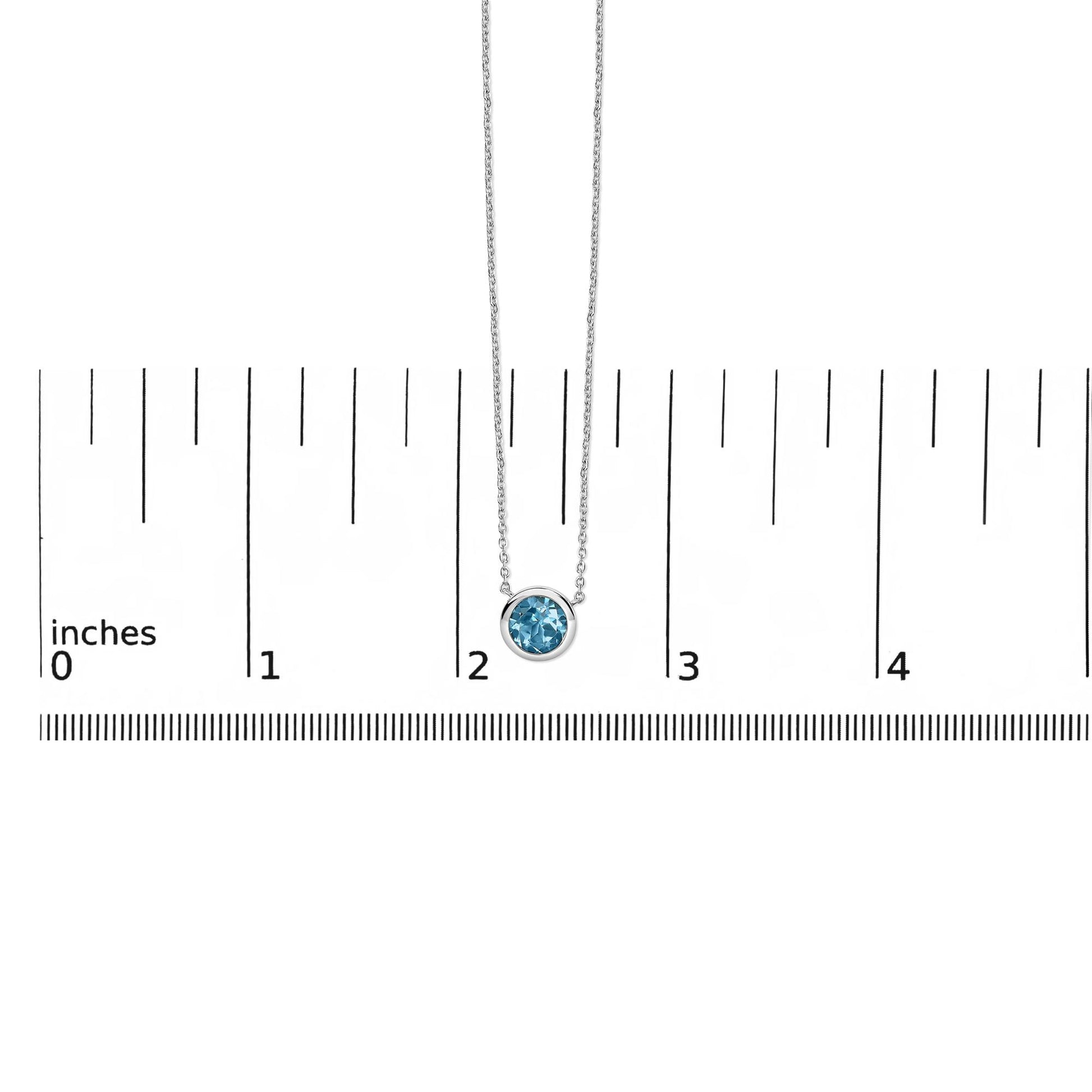 Close-up front view of a white necklace with a round blue gemstone in a bezel setting, shown on a measuring scale.