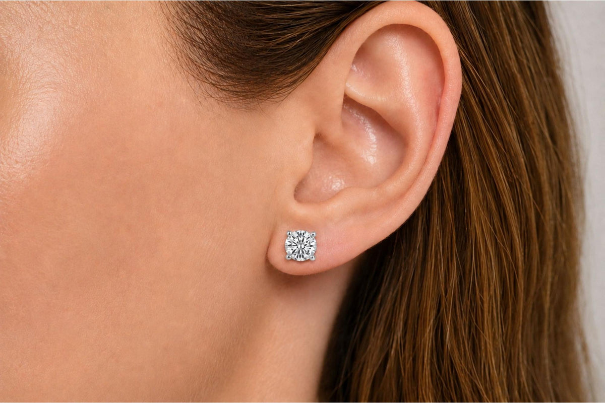 Close-up side view of a model's ear wearing a white metal earring with a round white gemstone in a four-prong setting.