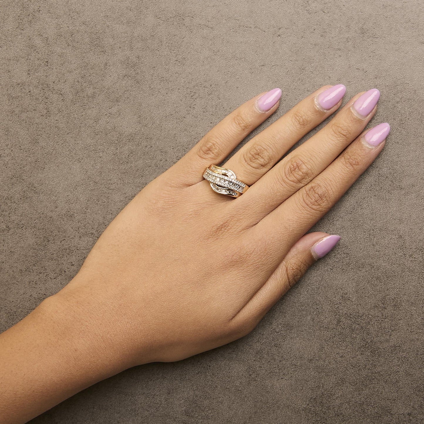 Yellow metal ring with intertwined bands featuring round white gemstones in prong settings and baguette white gemstones in channel settings, shown worn on a model's hand from an overhead angle.
