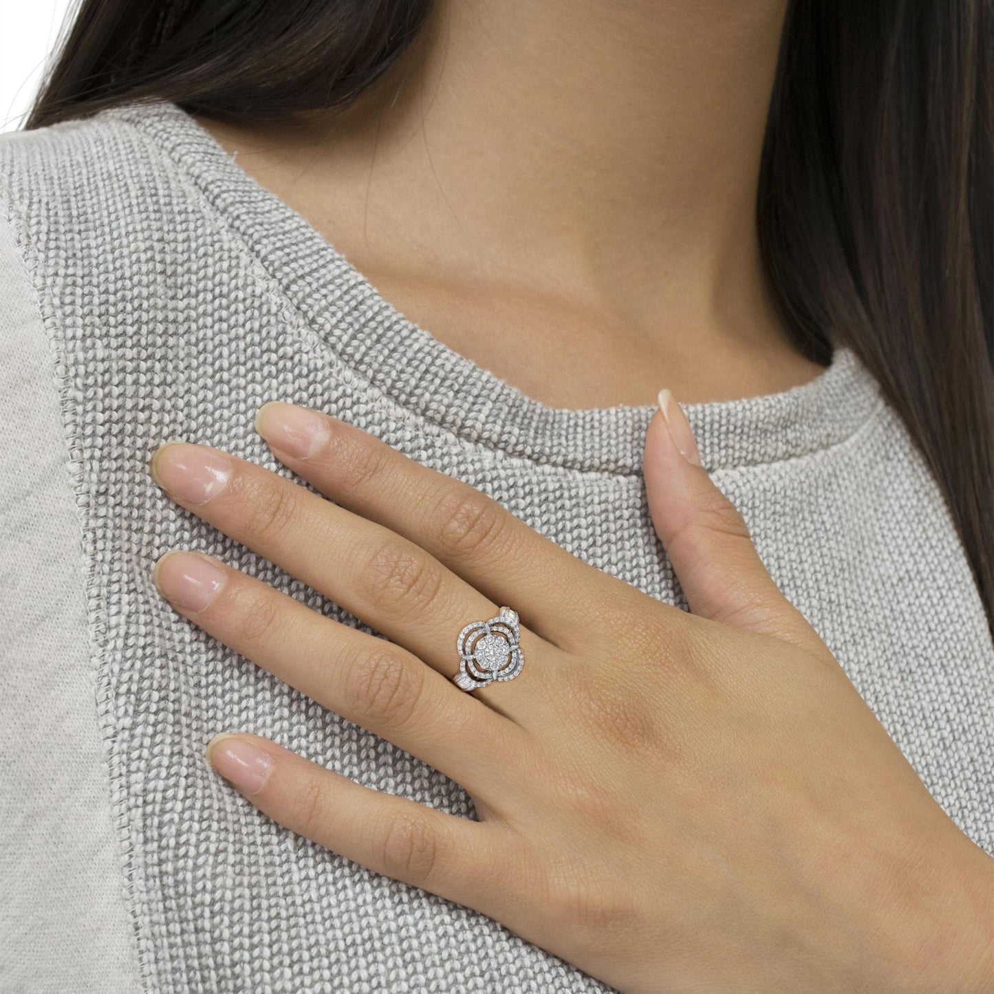 Close-up angled shot of a white metal ring with a round gemstone centerpiece, surrounded by smaller round and baguette-shaped gemstones, worn on a model's finger against a gray textured sweater.