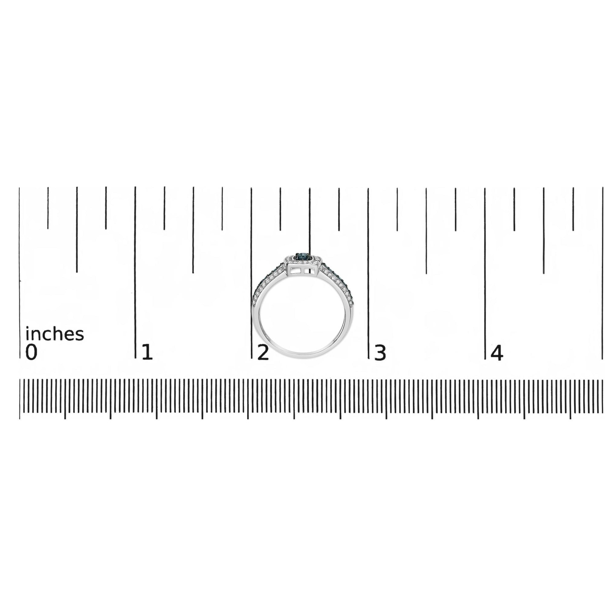 White metal ring with a single round blue gemstone center, flanked by multiple smaller round blue and white gemstones along the band, prong settings, positioned upright on a ruler for size reference, front angle.