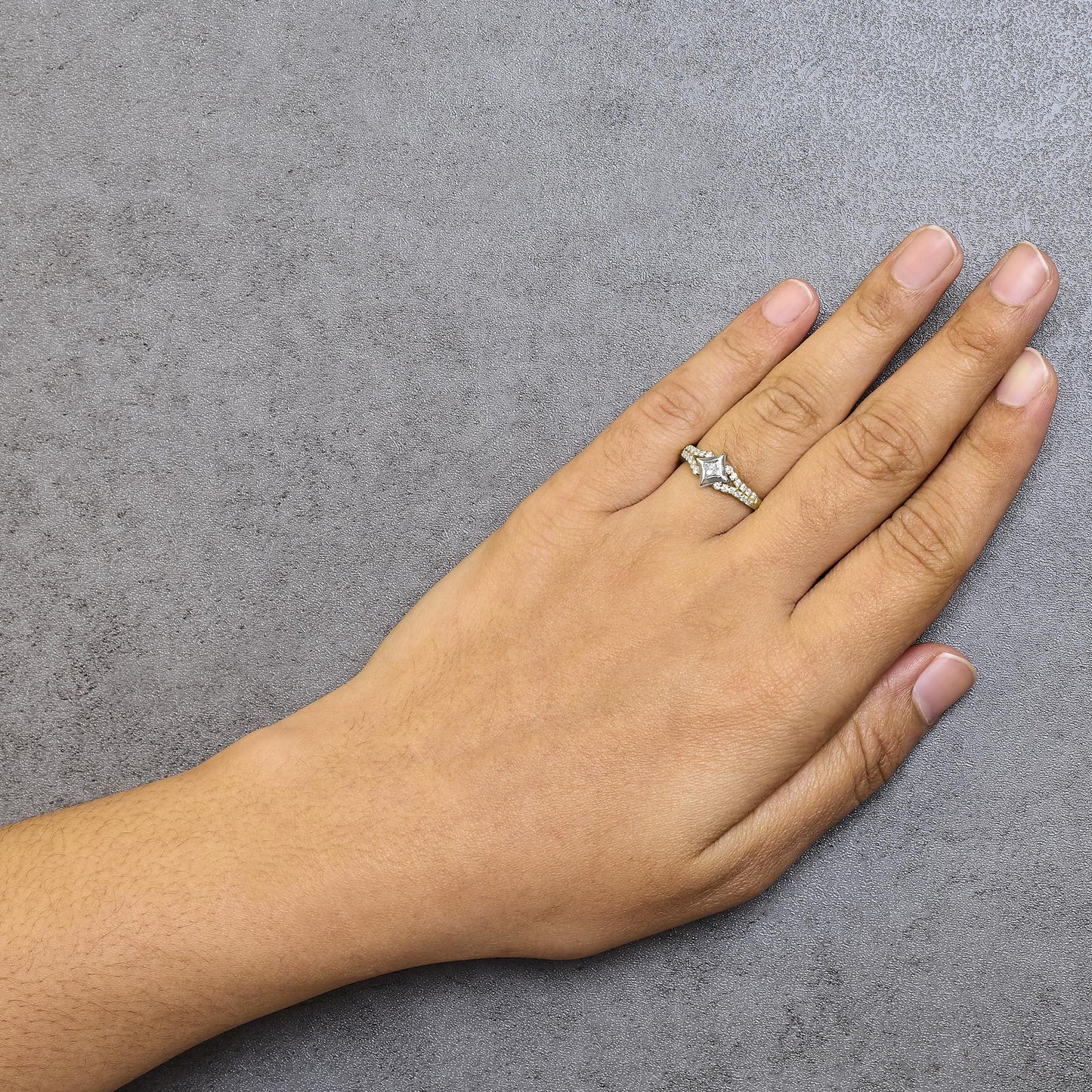 Ring with yellow and white metal featuring a princess shape gemstone in channel setting and multiple round shape gemstones in prong settings, worn on a model's finger, top-down angle.