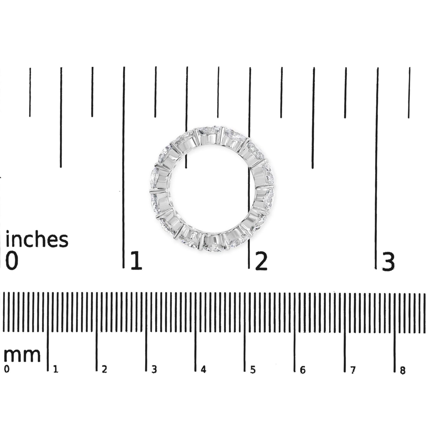 Top-down view of a white metal ring with round white gemstones in a shared prong setting, placed on a ruler for size reference.