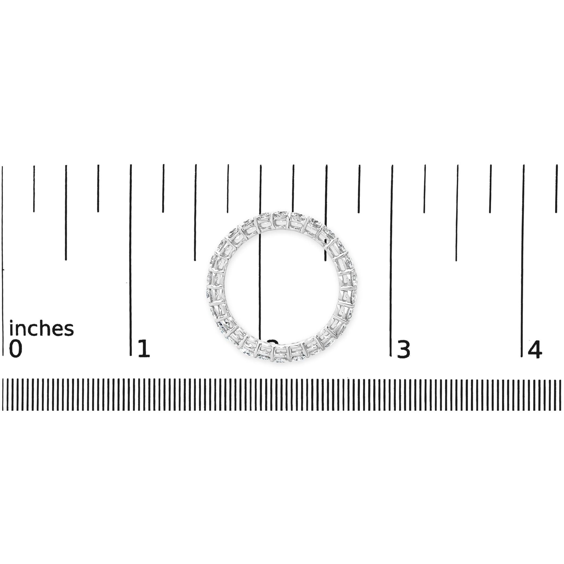 Top view of a white metal ring with round white gemstones in prong settings encircling the band, placed on a ruler for size reference.