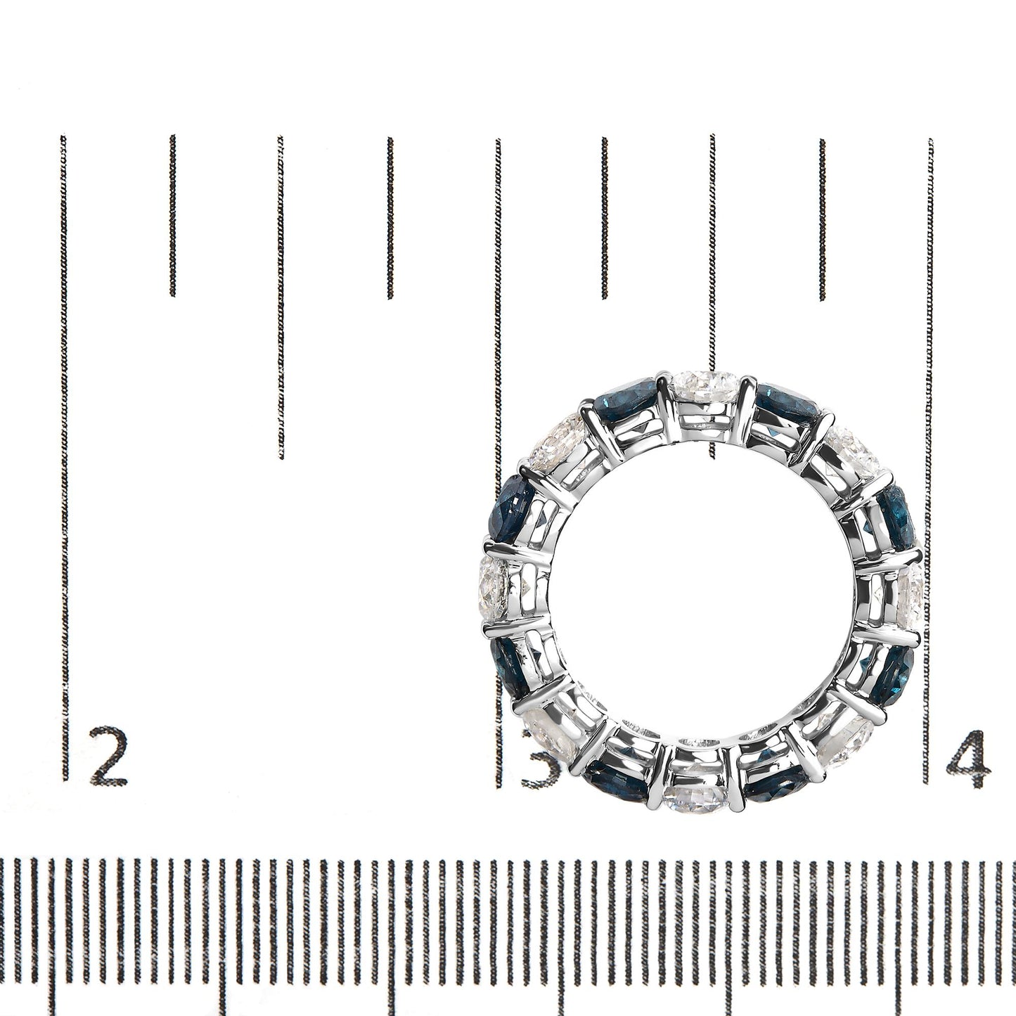 Top-down view of white metal ring with alternating round blue and white gemstones in shared prong setting, placed on a ruler for size reference.