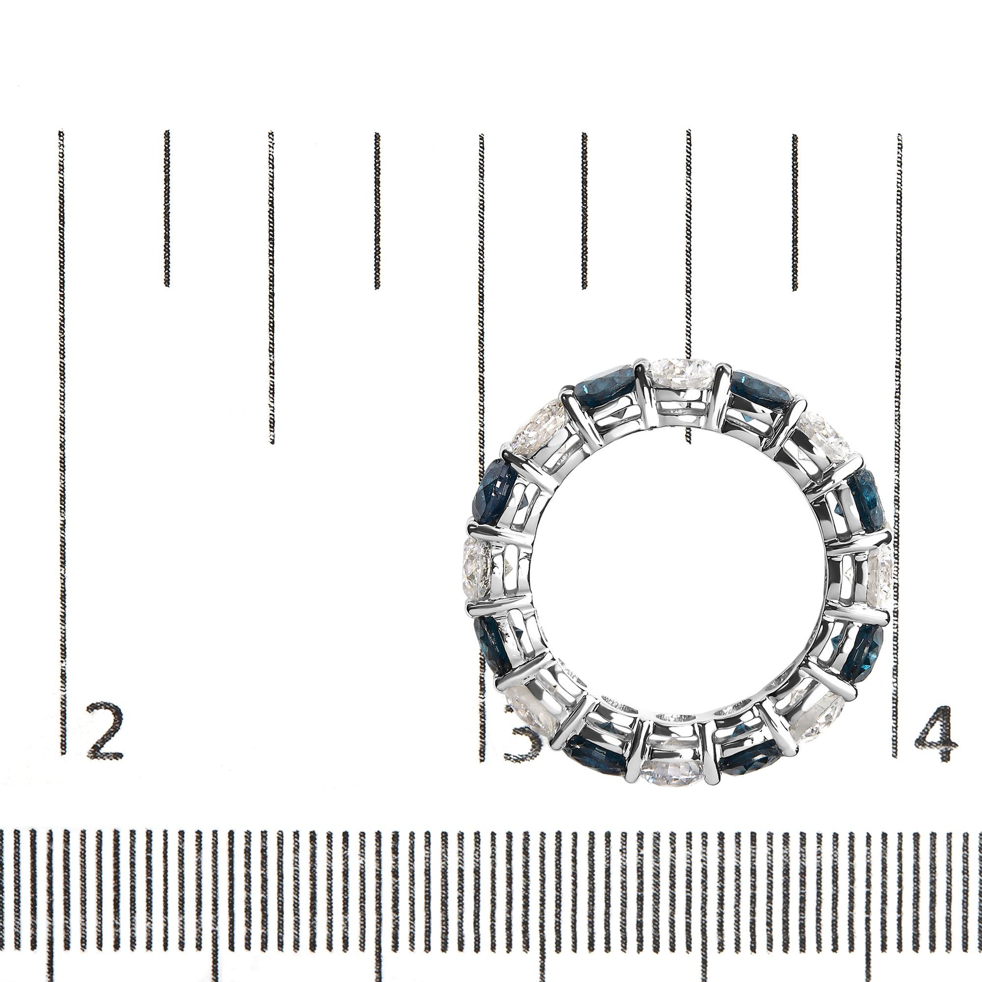 Top-down view of white metal ring with alternating round blue and white gemstones in shared prong setting, placed on a ruler for size reference.