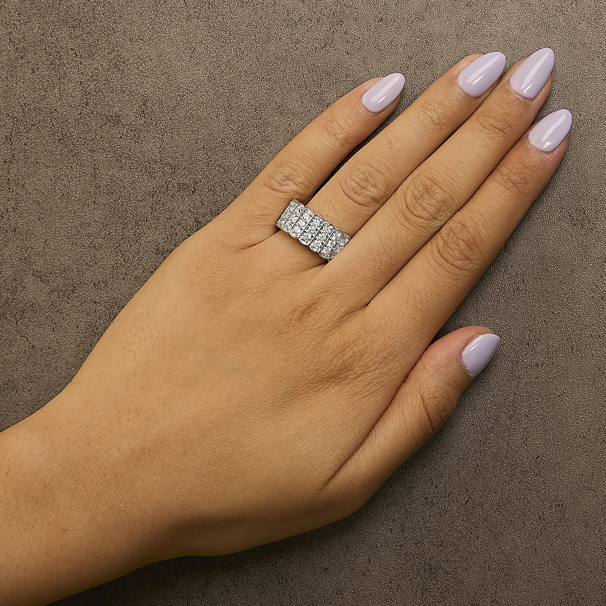 Close-up front angle of a white metal ring with multiple rows of round white gemstones in prong settings, shown worn on a model's ring finger against a textured background.