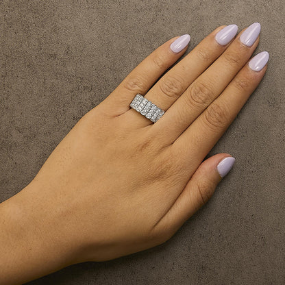 Close-up front angle of a white metal ring with multiple rows of round white gemstones in prong settings, shown worn on a model's ring finger against a textured background.