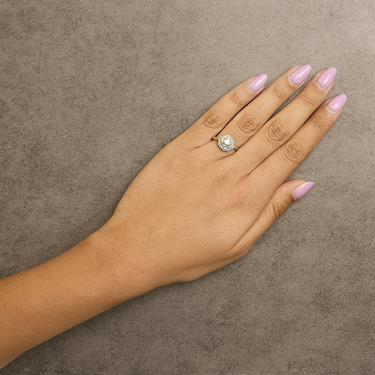 Yellow metal ring with a round white gemstone in a 4-prong setting, designed with a flower shape around the gemstone, shown on a model’s hand from a top angle.
