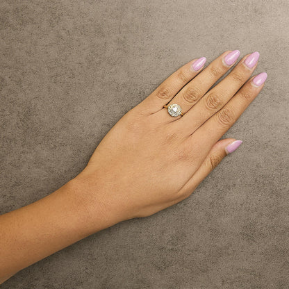 Yellow metal ring with a round white gemstone in a 4-prong setting, designed with a flower shape around the gemstone, shown on a model’s hand from a top angle.